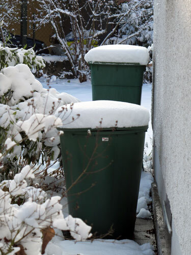 Zwei grüne Regentonnen mit Schneehaube an der Hauswand im Winter
