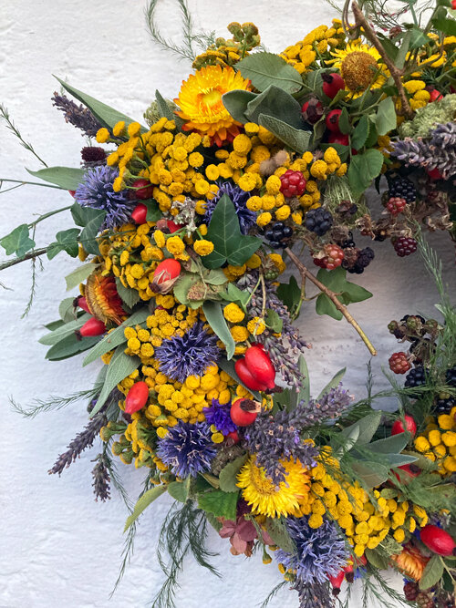 Ausschnitt eines fertigen Herbstkranzes mit gelben und blauen Blüten, roten Hagebutten und dunklen Beeren
